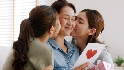 Wall Mural - Mother day two grown up child cuddle hug give flower gift box red heart card to mature mum. Love kiss care mom asia middle age adult three people sitting at home sofa happy smile enjoy family time.