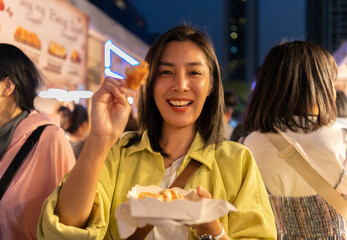 Wall Mural - Asian woman enjoy eating fries street food at night market. Traveler Asian blogger women Happy tourists Beautiful female with Traditional thailand bangkok food.