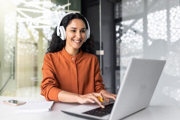 Wall Mural - Young successful hispanic woman working in office with headphones, female programmer coding software on laptop smiling, listening to online audio books and podcasts, businesswoman inside office.