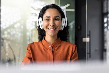 Young successful hispanic woman working in office with headphones, female programmer coding software on laptop smiling, listening to online audio books and podcasts, businesswoman inside office.