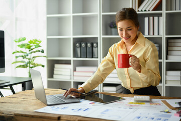 Wall Mural - Cheerful asian woman employee drinking coffee and working with statistic reports at office desk