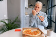 © dikushin - front view of handsome hungry bearded mature elderly male enjoying to eat pizza alone sitting at kitchen table. Front view of bearded senior older man with appetite to eat tasty pizza at home.