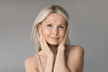 Aged skincare concept. Beautiful senior lady touching her face and smiling, posing on grey wall background