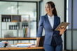 © crizzystudio - Asian businesswoman in the suit standing confidently holding iPad or tablet in hand and smiling in the office.