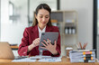 © crizzystudio - Asian businesswoman in a suit sitting holding iPad or tablet in contact See details and discuss business information happily in the office.