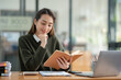 © crizzystudio - Beautiful Asian businesswoman working on a laptop and having fun at work and reading review book documenting detailed financial information earning peace of mind.