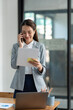 © crizzystudio - Attractive Asian businesswoman standing on the phone talking about real estate projects and chatting with happy smiles at the office.