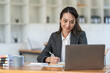 © crizzystudio - Young Asian businesswoman holding a pen sitting and analyzing the details of management information Finance is happy at the desk in the office.