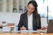© crizzystudio - Asian businesswoman working at office pressing calculator to calculate and writing company account on graph paper. Review various information and management concepts.
