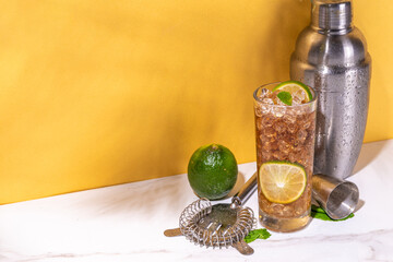  Cuba libre or long island summer cocktail, iced cola with lime and mint. Iced sweet long alcohol drink with crushed ice and bar utensils on high-colored golden beige background 