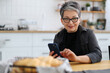 © Andrii Zastrozhnov - Smiling senior woman with a phone in her hands sits at a table against the backdrop of a home kitchen.