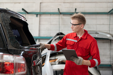 Car mechanic man in red uniform using tablet checking rear window broken of car at garage auto service. Car repair and maintenance concept.