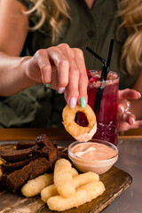 Wall Mural - Girl dipping fried onion rings in sauce, snacks rye bread croutons, crispy onion rings, cheese sticks and sauce.