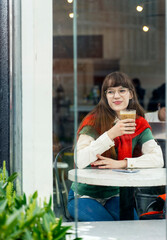 Positive, beautiful woman wearing glasses, drinking coffee, sitting in modern cafe looking at window