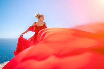 woman sea red dress. Blonde with long hair on a sunny seashore in a red flowing dress, back view, silk fabric waving in the wind. Against the backdrop of the blue sky and mountains on the seashore.