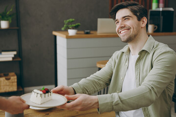 Wall Mural - Young smiling happy fun caucasian man wear shirt casual clothes receive order take cake from waiter sitting alone at table in coffee shop cafe restaurant indoors rest relax during free time inside.