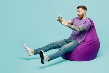 Full body gambling young man in purple t-shirt sit in bag chair use play racing app on mobile cell phone hold gadget smartphone for pc video games isolated on plain pastel light blue cyan background