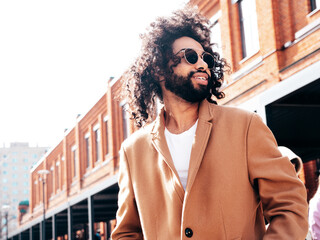 Handsome smiling hipster model.  Unshaven Arabian man dressed in brown coat clothes. Fashion male with long curly hairstyle posing outdoors in street. Cheerful and happy. In sunglasses. At sunny day