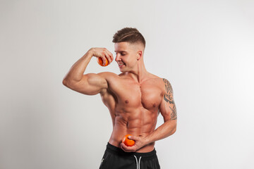Happy handsome fitness trainer man with a muscular body with oranges shows biceps on a white background in the studio. Healthy food, exercise and vitamins