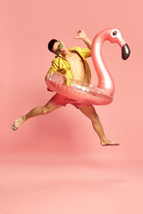 Portrait with attractive young guy, man wearing beach clothes jumping with inflatable flamingo over pink studio background. Pool party