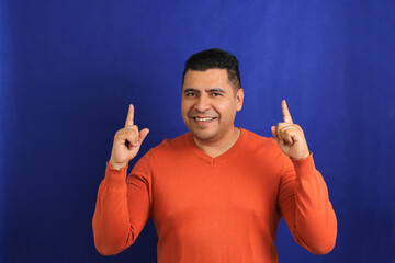 Adult man forty years old latino brunette with orange sweater points with his hands blue background copy space with happy and natural expression