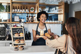Happy woman, barista and serving customer at cafe for service, payment or order on counter at coffee shop. African person, waitress or employee in small business restaurant helping client at checkout