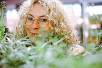 Portrait of happy blonde in glasses with curly luxurious hair in afternoon in garden center among plants. Rest and pleasure. Unpretentious plants for the winter garden. Hobby. Positive emotions.