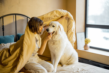 Cute young woman playing with her white adorable dog while sitting together covered with blanket on bed at home. Concept of friendship with pets and home coziness