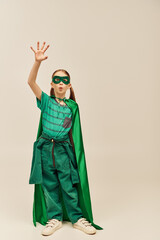 surprised girl in green superhero costume with cloak and mask on face, pouting lips and standing with outstretched hand while celebrating Worlds Child protection day holiday on grey background