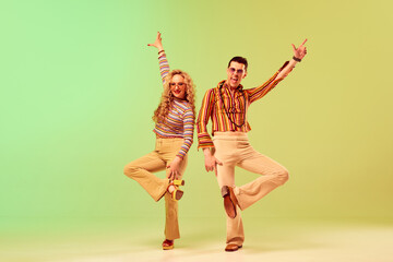 Expressive, talented young couple, man and woman in stylish vintage costumes dancing against gradient green yellow background. Concept of retro style, disco dance, fashion, art, hobby, music, 70s