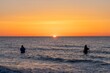 Die Silhouetten von zwei  Anglern in wasserdichten Hosen angelen vor einem traumhaften orangen Sonnenuntergang  in der Ostsee. Sie stehen mitten im Wasser der Ostsee.
