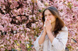 © sakoqwakestock - Sick Young Woman with Dark Hair Sneezing in Napkin While Standing Near Blooming Sakura Tree on City Street. Symptoms from Seasonal Allergy