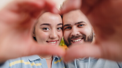 Young family married couple man woman makes symbol of love, showing heart sign to camera, express romantic feelings, express sincere positive feelings at home. Husband, wife together on sofa in room