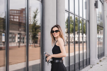 Wall Mural - Attractive beauty woman walking on the street while wearing black tank top, pants and glasses, mobile phone on tape . Blonde girl is turn around near office building. Mirror windows.