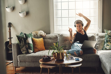 Dancing, headphones and woman on sofa and laptop, happy energy and mental health music in her home. Young person on couch listening to audio tech, electronics and podcast on computer in living room