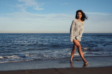 Wall Mural - young beautiful brunette girl walks on the sand on the seashore in a swimsuit and a sweater