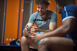 © luckybusiness - Young man using mobile phone with friend sitting on bench in the gym locker room, having fun before workout.