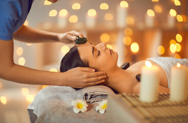 Wall Mural - Pretty woman is getting face massage at spa salon. Cosmetologist is doing facial massage with jade roller to relaxed young woman lying on towel with flowers on massage bed. Beauty, skin care concept