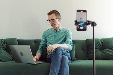 Wall Mural - Man in a T-shirt and jeans is sitting at home on couch in front of phone camera and typing something in notebook. Modern technologies. Online training. Male blogger. Video chat.