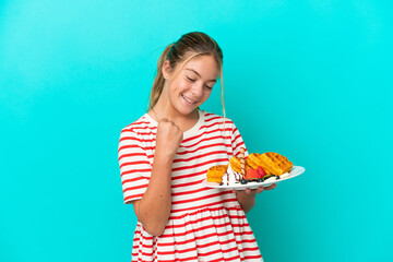 Wall Mural - Little caucasian girl holding waffles isolated on blue background celebrating a victory