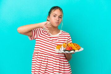 Wall Mural - Little caucasian girl holding waffles isolated on blue background showing thumb down with negative expression