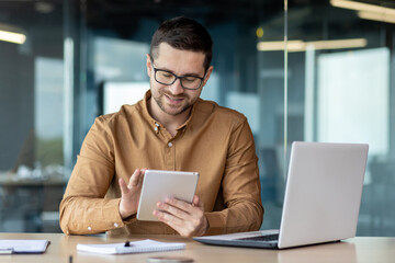 Portrait of a smiling young man designer, engineer, architect sitting in the office behind a notebook and works, draws, doodles online on the tablet he is holding in his hands