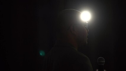 Canvas Print - 
Confident successful speaker man talking on stage with spotlight strike through the darkness at corporate business event. Public speaker giving talk at conference hall. Stand up comedian.
