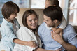 Cheerful cute little kids hugging positive mom and dad on sofa, smiling, laughing. Happy couple of parents playing with children on sofa, enjoying care, family leisure time, playtime