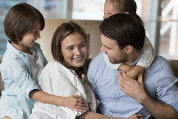 Wall Mural - Cheerful cute little kids hugging positive mom and dad on sofa, smiling, laughing. Happy couple of parents playing with children on sofa, enjoying care, family leisure time, playtime