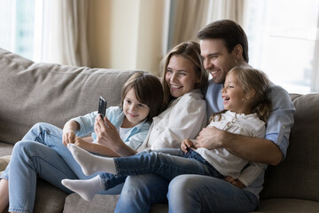 Wall Mural - Young mother and father hugging cheerful kids on home sofa, taking happy family picture on smartphone, using mobile phone together, for online communication, enjoying domestic Internet technology