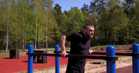 A man in the park at the training ground does push-ups on the uneven bars. A handsome man goes in for sports in the summer outdoors on a sports ground. Sports man concept.