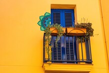 House With A Bright Yellow Exterior, Blue Windows And Balcony Adorned With Flowers To Bring Life To The Building.