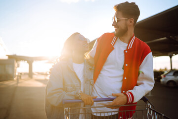 Wall Mural - Stylish man and woman having fun and riding shopping cart. Black friday. Consumerism, sale, discounts, lifestyle concept.