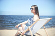© rogerphoto - Happy young brunette woman relaxing on a wooden deck chair at the ocean beach while smiling, and wearing fashion sunglasses. The enjoying vacation concept
